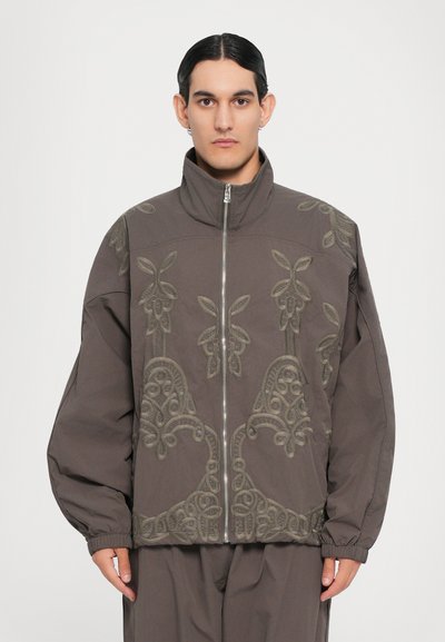 Chaqueta gris con alto cuello y cierre de cremallera. Presenta un intrincado bordado verde y mangas de gran tamaño, hecha de un tejido suave.
