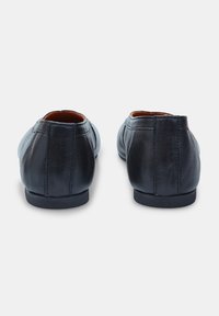 Mocasines de cuero negro con una textura suave, parte posterior redondeada y pequeñas suelas de goma. Presentan costuras cosidas y un diseño minimalista.