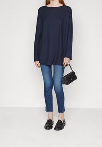 Pull oversize bleu marine, jean skinny bleu clair, mocassins noirs. Le modèle tient un petit sac à main noir au design structuré. Fond simple.