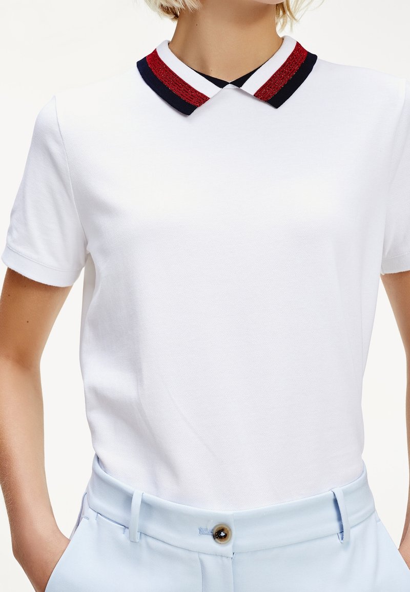 Personne portant un polo blanc à manches courtes avec un col à rayures noires et rouges, associé à un pantalon bleu clair.