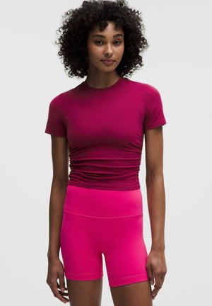 Vrouw met krullend haar die een nauwsluitend magentarood kort mouwen topje en felroze sportshort met hoge taille draagt, staand tegen een effen achtergrond.