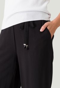 Zwarte broek met een tailleband met trekkoord, voorzien van gevlochten koord en metalen accenten, naast een witte top met een gladde textuur.