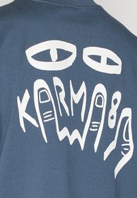 Blue fabric t-shirt with a large white graphic featuring eyes and abstract hand shapes, displaying the word "KARMA" in stylized text.