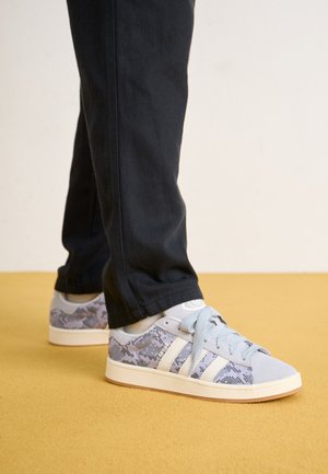 Person iført lyseblå og grå sneakers med slangeskindsprint og sorte bukser, der står på et gult gulv.