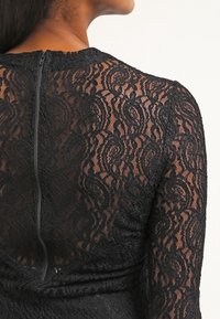 Vue arrière d'une personne portant un top en dentelle noire ajusté avec une fermeture éclair visible dans le dos et des motifs floraux complexes.
