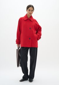 Rode wollen jas met een kraag, twee knopen en zijzakken. Gecombineerd met zwarte wijde broek en een bruine leren tas. Gladde textuur.
