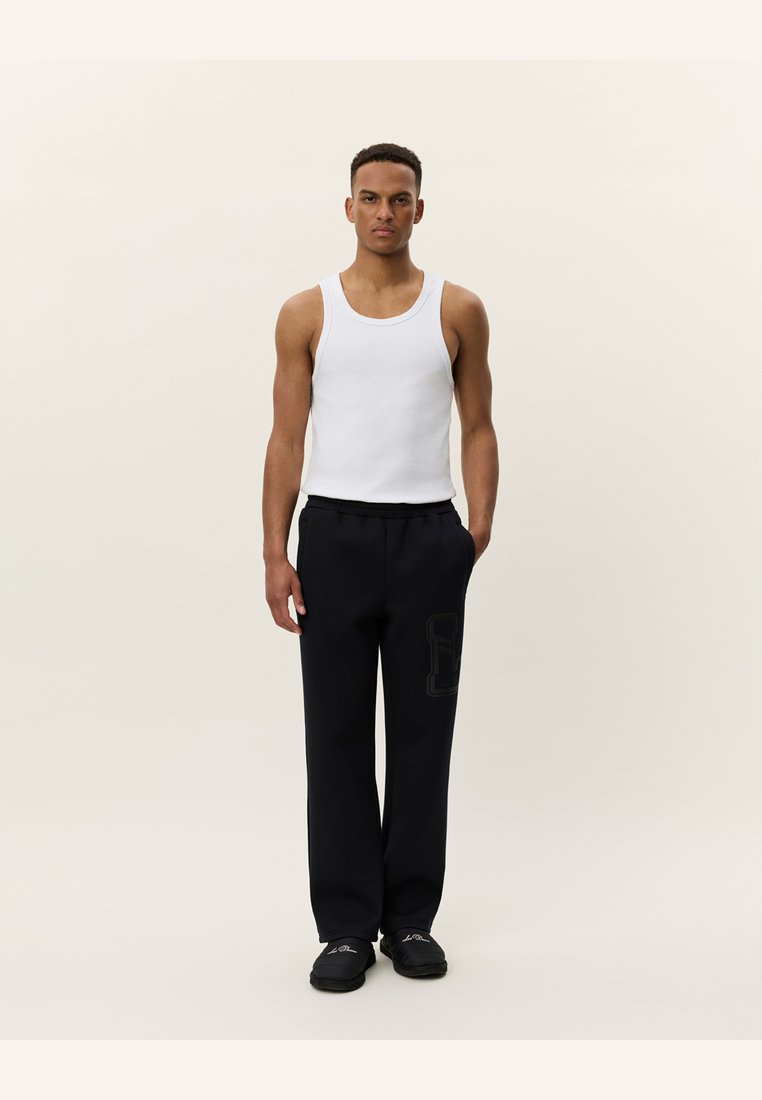 Hvid bomulds tanktop, sorte afslappede sweatpants med sidelomme-detalje og sorte slip-on sko. Enkelt baggrund.