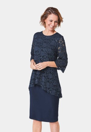 Marineblaues Kleid mit einer Spitzenübergardine, die Blumenmuster zeigt, dreiviertel langen Ärmeln und einem asymmetrischen Saum. Glattes, elastisches Unterkleid.