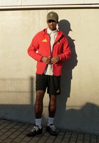 Raudona sportinė striukė su geltonomis detalėmis, balta marškinėliai, juodi šortai, baltos kojinės ir juodi sportbačiai. Fone – pilka siena.