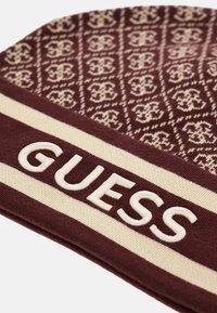 burgundin värinen neulepipo, jossa on vaalea geometrinen kuvio ja leveä vaalea reunus, jossa on brodeerattu "GUESS" lihavoiduin kirjaimin.