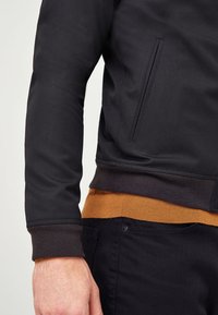 Zwarte jas met ritssluiting en een gestructureerde afwerking, voorzien van een bruine geribbelde zoom en manchetten. Heeft een onopvallende zijzak.