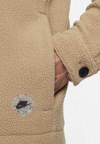 Beige fleecová bunda s texturovanou tkaninou, boční kapsou, černým knoflíkem a jasným logem Nike s barevnými skvrnami.