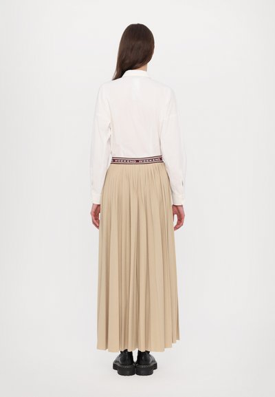 Chemise blanche à manches longues avec col, jupe plissée beige, et bottes noires à semelles épaisses. La taille élastique présente l'inscription "WEEKEND" imprimée en rouge.