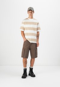 Striped short-sleeve beige and white t-shirt paired with brown cargo shorts; model wears black lace-up boots and a gray cap.