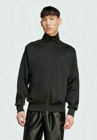 Ung mann iført svart Adidas treningsjakke og skinnende svarte bukser, stående mot en enkel lys bakgrunn.