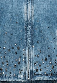 Τζιν μπουφάν με ανοιχτό μπλε ύφασμα διακοσμημένο με διάφορα μαύρα τρουκς και στρας, τοποθετημένα σε διάσπαρτο μοτίβο κατά μήκος του τελειώματος.