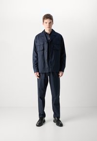 Navy blå jakke med to frontlommer, knappelukking og matchende bukser. Glatt tekstur og avslappet passform, kombinert med svarte sko.