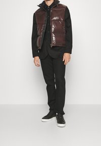 Braune glänzende Steppweste mit Reißverschluss, getragen über einem schwarzen Hemd mit Knöpfen und schwarzen Hosen. Schwarze Sneakers runden den Look ab.