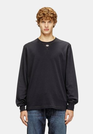 Giovane uomo con capelli ricci che indossa una felpa nera semplice a maniche lunghe e jeans blu, in piedi davanti a uno sfondo grigio chiaro.