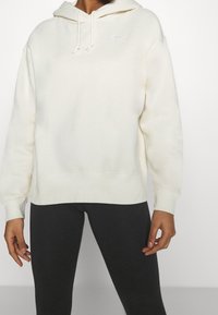 Felpa bianca realizzata in tessuto morbido, dotata di cappuccio, cordini e un piccolo logo sul petto. Abbinata a leggings neri.