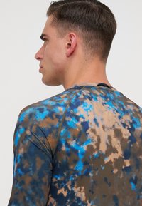 Cămașă cu mâneci lungi din material elastic, cu un model tie-dye în nuanțe de brun și albastru. Textură netedă cu guler rotund și cusături în contrast.