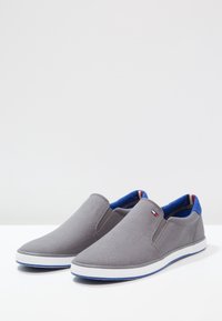 Scarpe slip-on in tela grigia con rivestimento interno blu, suola in gomma bianca e un piccolo logo sul lato. Punta rotonda e design minimalista.