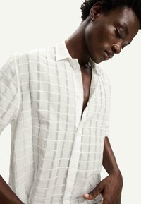 Camicia bianca con bottoni realizzata in tessuto leggero, caratterizzata da un motivo a griglia con linee grigie, maniche corte e una vestibilità rilassata.