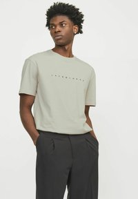 Helles graues Kurzarm-T-Shirt mit einem dezenten schwarzen Logo "JACK & JONES", das mittig auf der Brust platziert ist, kombiniert mit dunklen Hosen.