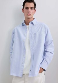 Camicia a righe blu chiaro con bottoni e tasca, indossata sopra una t-shirt bianca, caratterizzata da maniche lunghe e una vestibilità comoda.