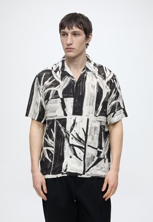 Homme portant une chemise à manches courtes boutonnée à motif abstrait noir et blanc avec un pantalon noir, debout sur un fond blanc.