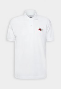 Hvit polo shirt laget av bomull, med krage med to knapper, korte ermer og en rød brodert krokodillelogo på venstre bryst.