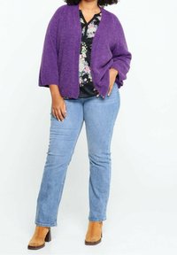 Cardigan viola oversize con maniche lunghe, texture morbida; indossato sopra una camicetta floreale e abbinato a jeans azzurri e stivaletti color cuoio.