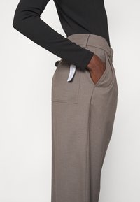 Ljusgrå skräddarsydda byxor har en vid benvidd och framfickor, med en platt midja och en märkning synlig bak. Slät tygtextur.