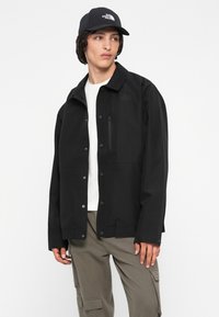 Mladenič oblečen v črno jakno The North Face in kapo, belo majico in bež cargo hlače, gleda levo na enobarvni beli ozadju.