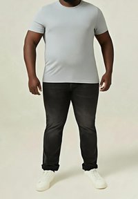 Homme portant un t-shirt ajusté gris clair, un jean noir avec les ourlets retroussés et des baskets blanches, debout devant un fond clair uni.