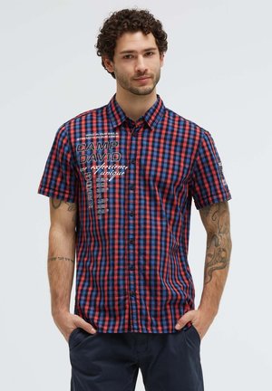 Man with curly hair wearing a red and blue checkered short-sleeve shirt with text and navy pants, hands in pockets, neutral background.