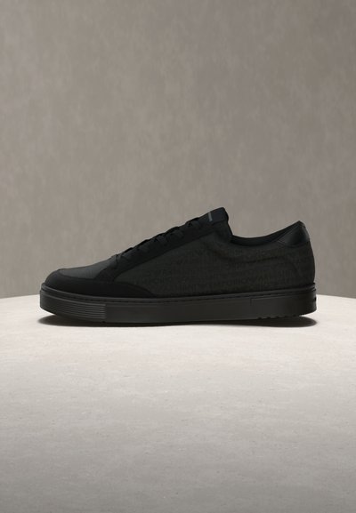 Μαύρο αθλητικό παπούτσι με χαμηλό κόψιμο, διακριτικό μοτίβο με λογότυπο, μαύρα κορδόνια και λαστιχένια σόλα πάνω σε μια ελαφριά στρογγυλή επιφάνεια σε απλό γκρι φόντο.