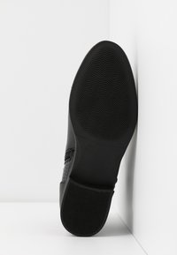 Semelle de chaussure noire avec des motifs de bande de roulement ondulés et droits, positionnée verticalement contre un fond blanc en coin.