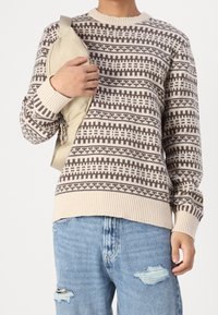 Beige tröja med bruna geometriska mönster, ribbat nacke och ärmslut; som bärs med slitna blå jeans och en beige axelväska.