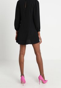 Schwarzes Kleid mit langen Puffärmeln und einem Schlüsselloch-Detail am Rücken, kombiniert mit leuchtend pinkfarbenen High Heels. Glatter Stoff und kurze Länge.