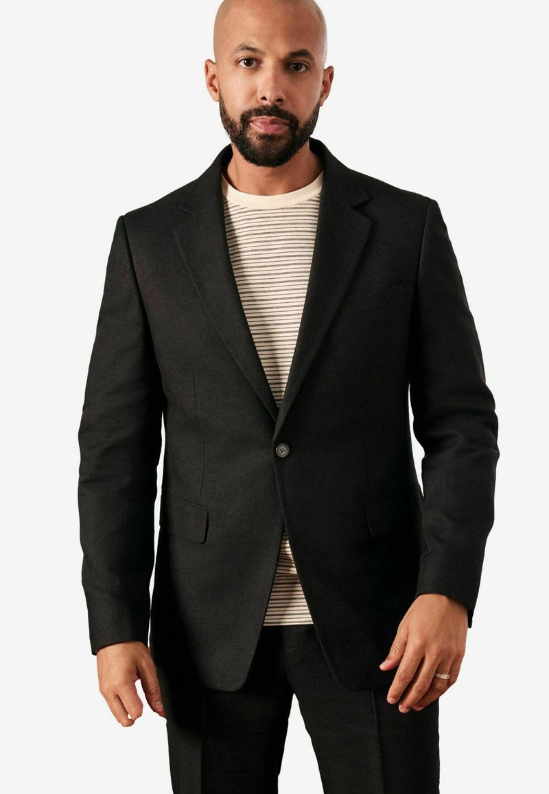 Blazer negro a medida con solapas con muesca, cierre de un solo botón y dos bolsillos delanteros, llevado sobre una camiseta de cuello redondo a rayas.