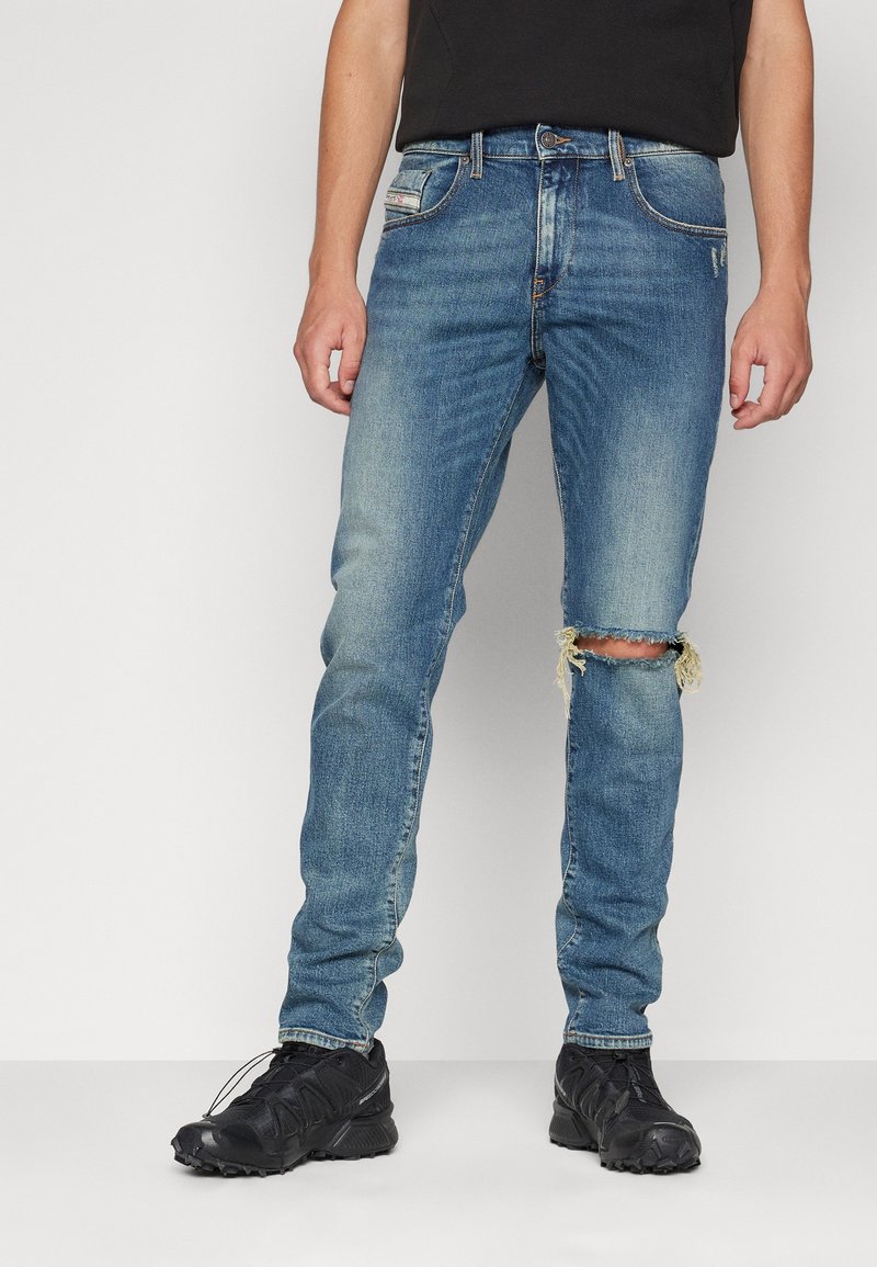 Blå denimjeans med slim fit, med ett slitet knä, blekt mönster och kontraststygn. Bärs med svarta sportskor.