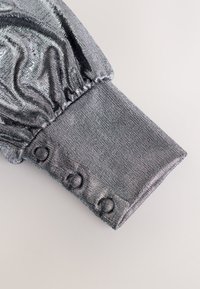 Manche en tissu argenté brillant avec un détail bouffant froncé et un poignet fermé par trois boutons recouverts de tissu.