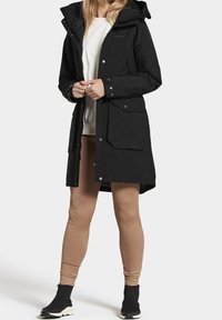 Personne portant un parka à capuche noir, des leggings côtelés beige, des baskets noires sans lacets et un sweat-shirt blanc sur un fond uni.