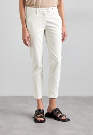 Mujer con camiseta beige, pantalones ajustados blancos hasta el tobillo y sandalias negras abiertas con doble hebilla sobre fondo gris.