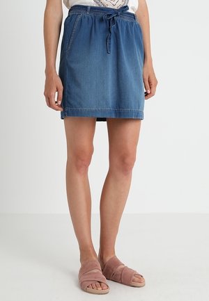 Femme portant une jupe en jean bleue avec une taille élastique et un cordon, associée à des sandales roses à enfiler, debout devant un fond uni.