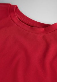 Prim-plan cu un tricou roșu de bumbac cu decolteu rotund, arătând textura țesăturii și detalii ale cusăturilor în jurul gulerului pe un fundal gri.