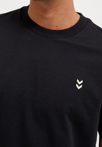 Gros plan d’une personne portant un t-shirt noir uni à col rond avec deux petits logos blancs en forme de chevrons sur la poitrine gauche.