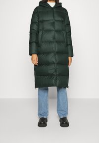 Person iført en lang, mørkegrøn puffjakke med sidelommer, lyseblå jeans og sorte chunky støvler, der står foran en ensfarvet baggrund.