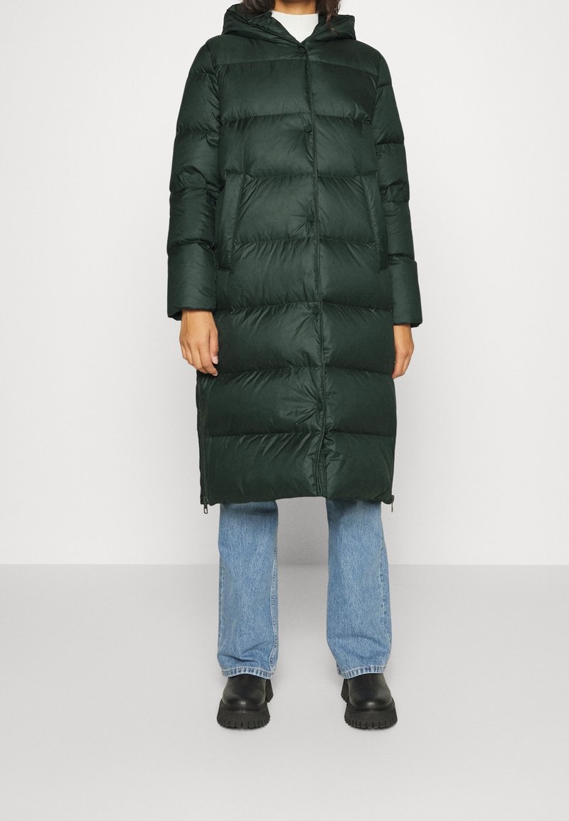 Person iført en lang, mørkegrøn puffjakke med sidelommer, lyseblå jeans og sorte chunky støvler, der står foran en ensfarvet baggrund.